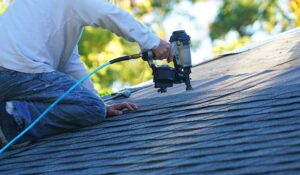 A person examining roofing shingles on a residential home with mature trees in the background.