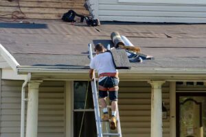 A person using a roof rake to remove snow from a shingled residential roof in cold, sunny weather.
