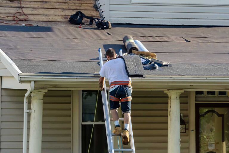 A person using a roof rake to remove snow from a shingled residential roof in cold, sunny weather.