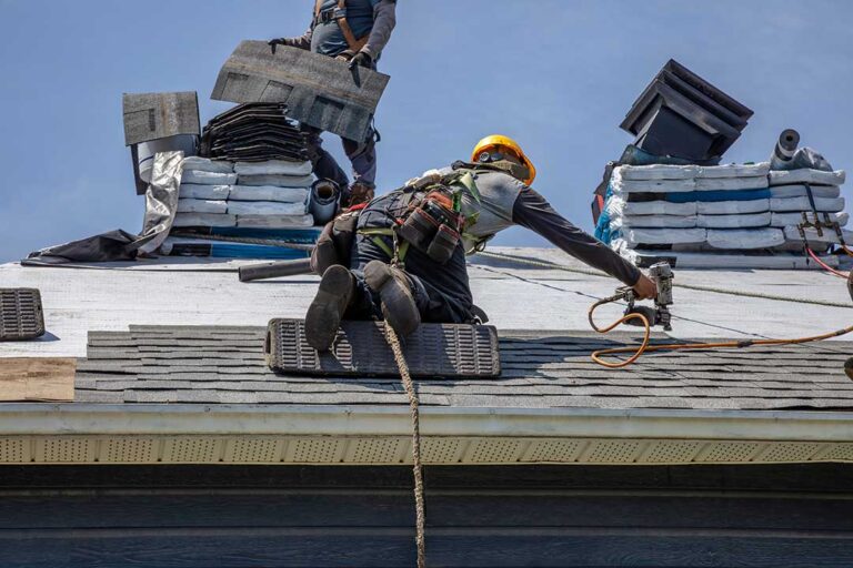 A pair of gloved hands cleaning leaves and debris from a residential rain gutter on a sloped roof.
